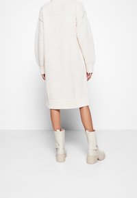 Robe oversize en maille couleur crème avec épaules tombantes et poignets côtelés, associée à des bottines beige à détails de sangles.