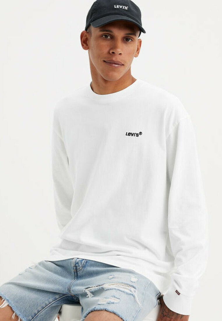 Uomo che indossa un cappellino nero Levi's, una maglietta bianca a maniche lunghe Levi's e pantaloncini in denim azzurro chiaro strappati, seduto davanti a uno sfondo semplice.