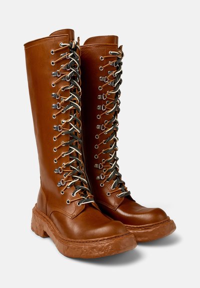 Botas de cuero marrón hasta la rodilla con una suela gruesa y texturizada, que presentan ojales de metal y cordones en colores contrastantes. Diseño de ojales secuencial.