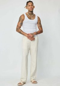 Débardeur blanc, pantalon beige ample et sandales. Tatouages notables sur les bras, debout avec les bras croisés. Fond clair.