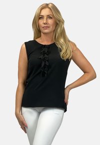 Czarny top bez rękawów z fakturowanym detalem falbany z przodu, wykonany z gładkiego materiału, w połączeniu z białymi dopasowanymi spodniami.