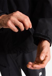Main ajustant la manche d'une veste noire avec une bande Velcro, montrant un gros plan des mains et d'une partie du vêtement sombre.