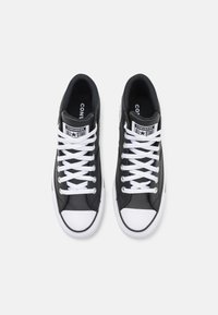 Adidași din piele neagră cu vârfuri și șireturi din cauciuc alb. Prezintă un logo proeminent pe limba încălțămintei și detalii cusute pe laterale.