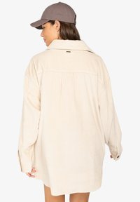Femme portant une grande veste en velours côtelé crème et une casquette de baseball grise, de dos, contre un fond blanc uni.