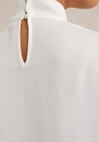 Vit blus med hög krage, knäppning bak och en liten nyckelhål detalj. Tillverkad av ett mjukt, texturerat tyg.