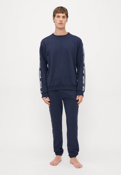Ung mann som står barbent i en navy blå sweatshirt og joggebukser med hvite logo-striper på ermene og sidene av buksebenene mot hvit bakgrunn.