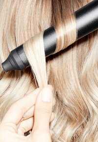 Hand die een blonde haarlok vasthoudt die om de cilinder van een zwarte krultang gewikkeld is, met gekruld haar op de achtergrond.