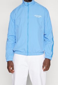 Chaqueta azul claro con cremallera y cuello alzado, con logotipos blancos y rayas laterales. Hecha de tela lisa con cinturilla elástica.