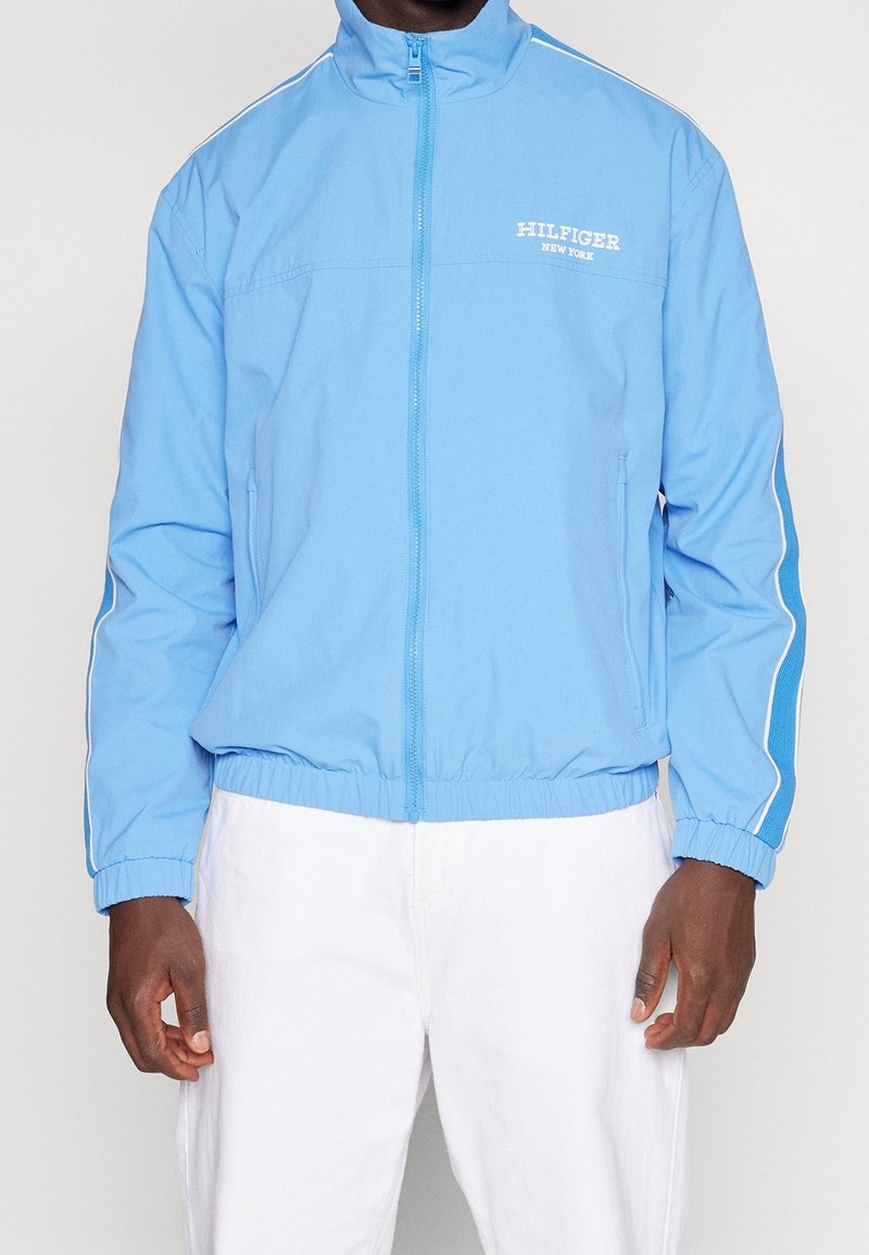 Chaqueta azul claro con cremallera y cuello alzado, con logotipos blancos y rayas laterales. Hecha de tela lisa con cinturilla elástica.