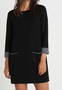 Vestito nero con maniche a tre quarti, polsini grigi e due tasche frontali. Realizzato in materiale morbido con un design semplice e lineare.