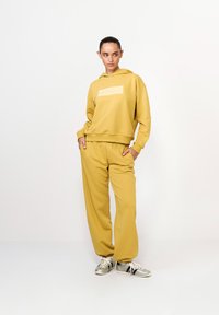 Žltá mikina s logom na prednej strane a zodpovedajúce žlté tepláky. Obidva kusy majú jemnú textúru; model má na sebe metalické tenisky.