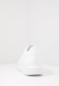 Vitt sneaker som står baklänges med tjock sula på en vit yta mot en vit vägg.