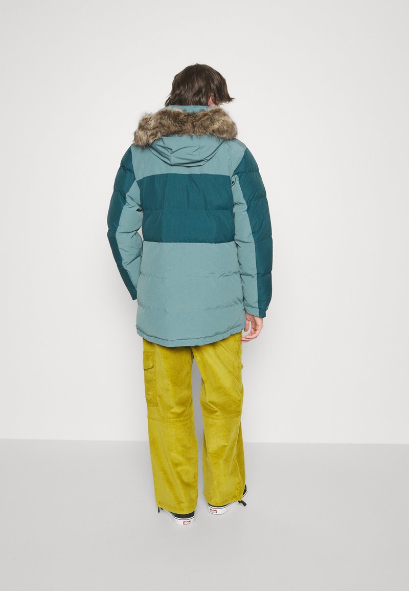 Veste matelassée en teal et vert foncé avec une capuche en fausse fourrure, assortie à un pantalon en velours côtelé jaune et des baskets noires. Fond blanc uni.