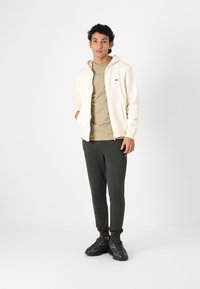 Creme farvet hættejakke med lynlås over en beige t-shirt, parret med mørkegrønne sweatpants og sorte sneakers, der har en lille logo-detalje.