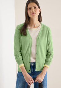 Jeune femme aux longs cheveux bruns portant un cardigan vert clair boutonné sur un pull léger et un jean bleu, tenant un objet noir.