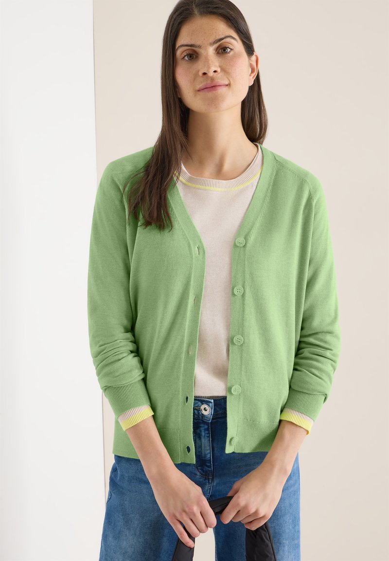 Jeune femme aux longs cheveux bruns portant un cardigan vert clair boutonné sur un pull léger et un jean bleu, tenant un objet noir.