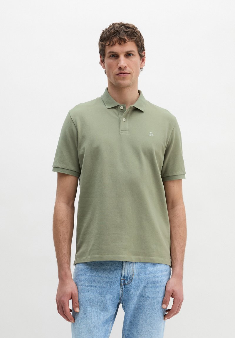 Homme debout devant un fond uni, portant un polo vert clair et un jean bleu clair, faisant face à l'avant avec une expression neutre.