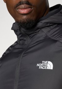 Svart nylon zip-up-jakke med høy krage, med en hvit logo på venstre bryst og en subtil teksturfinish.