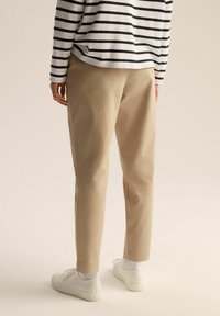 Beżowe spodnie taperowane o gładkiej fakturze, w połączeniu z pasiastym topem w granatowo-białe paski oraz białymi sneakersami. Skupienie na widoku z tyłu.