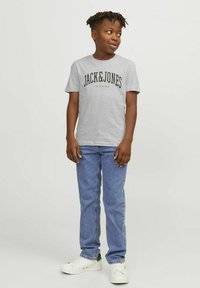 Camiseta gris de manga corta con gráfico negro "JACK & JONES", combinada con jeans de pierna recta azules y zapatillas blancas con acentos verdes.