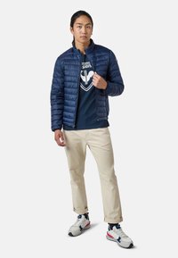 Mörkblå puffjacka med horisontell quiltning, bärs uppknäppt över en mörkblå t-shirt, matchad med ljusbeiga byxor och sportiga sneakers.