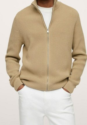 Pull en tricot beige à fermeture éclair porté sur une chemise blanche, associé à un pantalon blanc, mains dans les poches, coupe décontractée et confortable.