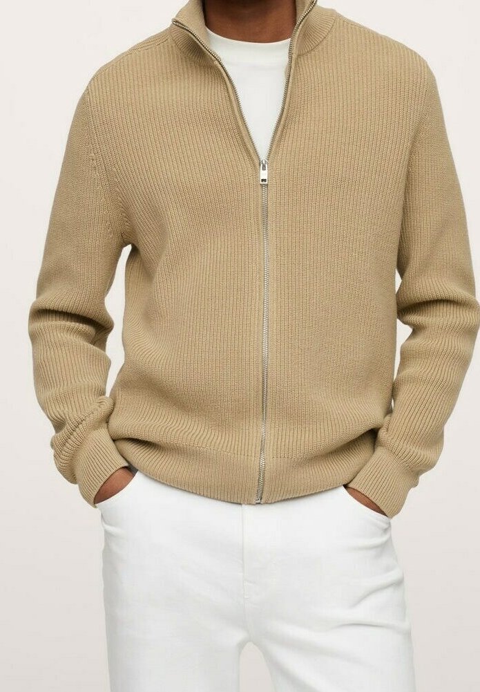 Pull en tricot beige à fermeture éclair porté sur une chemise blanche, associé à un pantalon blanc, mains dans les poches, coupe décontractée et confortable.