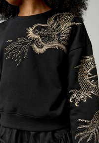 Sudadera negra con intrincados bordados de dragón dorado en el hombro y la manga izquierda, con un acabado texturizado y puños de canalé.