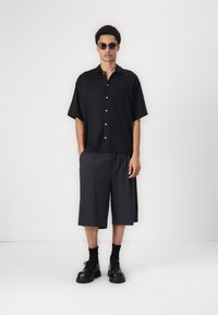 Svart kortermet skjorte med knapper, grå kne-lange shorts og svarte klumpete sko. Glatt stoff, avslappet passform og minimalistisk design.