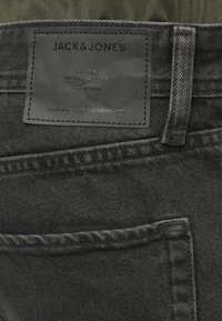 Juoda denimo džinsai su odiniu skydu, pažymėtu "JACK & JONES", virš galinio kišenės ir matomu diržo kilpos.