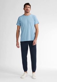 Lichtblauw katoenen T-shirt met korte mouwen, gecombineerd met marineblauwe joggerbroek. Model draagt witte sneakers en staat tegen een neutrale achtergrond.