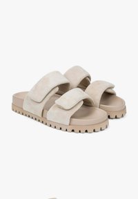 Slide sandals with two wide, soft, beige suede straps and a thick, tan rubber sole featuring a jagged texture for grip.