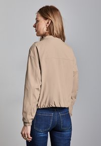 Světle béžová bomber bunda s elastickými manžetami a spodním lemem, zobrazená ze zadu. Nesená s tmavě modrými džíny a kruhovými naušnicemi. Hladká textura.