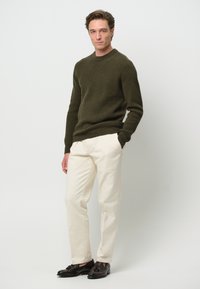 Olivgrön ribbstickad tröja, krämfärgade skräddarsydda byxor och mörka dressade skor. Enkla linjer och texturerade tygdetaljer.