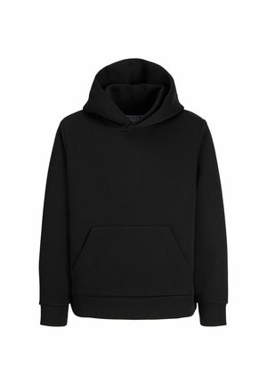Garçon blond debout et souriant portant un sweat à capuche noir, une chemise blanche en dessous, un jean gris ample et des baskets noires sur fond blanc.