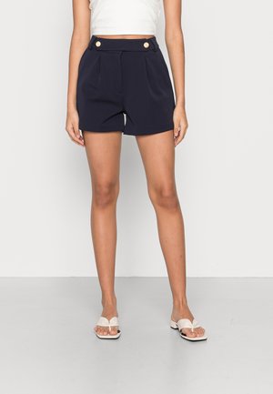 Kvinde iført marineblå shorts med høj talje, forrest plisserede detaljer og guldknapper, kombineret med hvide sandaletter med åben tå og hæl.
