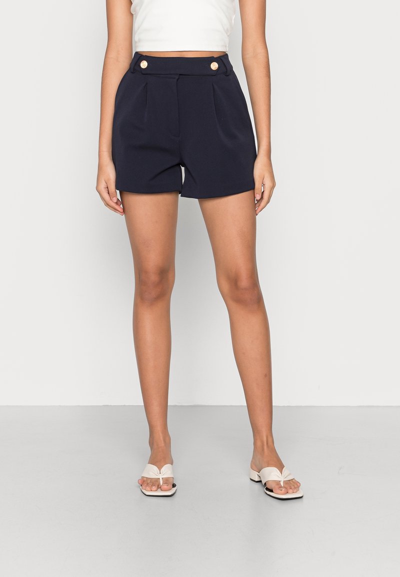 Femme portant un short taille haute bleu marine avec des plis sur le devant et des boutons dorés, associé à des sandales blanches à talons ouvertes.