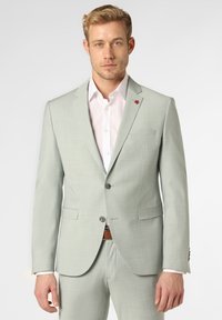 Hellgrüner Blazer mit strukturierter Oberfläche, zwei Knöpfen und einem kleinen roten Emblem auf dem Revers, kombiniert mit einem weißen Hemd und einem braunen Gürtel.