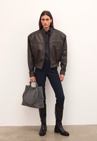 Nő oversized sötétbarna bőrkabátban, tengerészkék felsőben és nadrágban, fekete térdfelett érő csizmában áll egy nagy szürke táskával a fehér fal mellett.