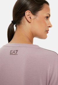 Světle fialová krátký rukáv košile s kulatým výstřihem. Na zadní straně je hnědý logo EA7 Emporio Armani. Hladká látka, minimalistický design.