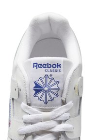 Fehér sportcipő párnázott nyelvvel, kék Reebok logóval, fehér cipőfűzőkkel, sima bőr felsőrészzel és textúrált sarokkal.