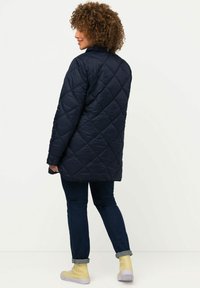 Námořnická prošívaná bunda s diamantovým vzorem, vysokým límcem a dlouhými rukávy. Nositá přes tmavé džíny a žluté kotníkové boty.