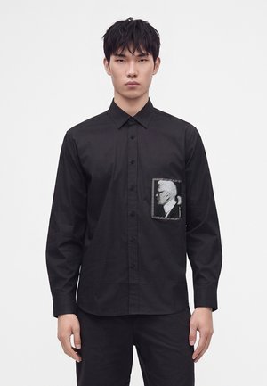 Giovane uomo che indossa una camicia nera con bottoni con una toppa rettangolare che mostra il profilo laterale monocromatico di una persona sulla tasca sinistra del petto.