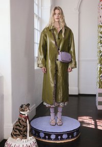 Zelený, lesklý, nadměrně velký kabát s černými knoflíky; přes květinové šaty. Modelka má na sobě žluté sandály na ramínka a drží malou levandulovou tašku.