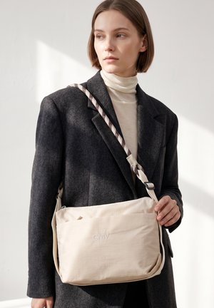 Vrouw met kort bruin haar, gekleed in een donkergrijze jas en een crèmekleurige coltrui, met een beige crossbodytas met het GMV-logo.