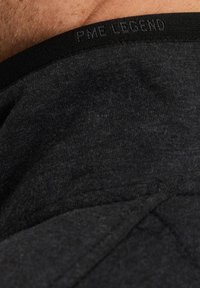Gros plan sur un tissu noir avec "PME LEGEND" brodé sur le bord, montrant la texture et une partie de la peau d'une personne au-dessus.