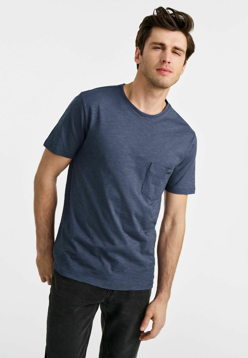 Granatowa koszulka z krótkim rękawem i okrągłym dekoltem, z małą kieszonką na piersi, wykonana z teksturowanej bawełny. Luźny krój z prostym dołem.