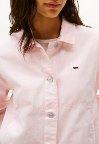 Giacca di jeans rosa con bottoni a pressione argentati, colletto classico ed emblema sul lato sinistro del petto. Tessuto testurizzato con cuciture visibili.