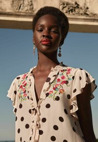 Blusa crema con pois marroni, maniche a ruches e ricami floreali in rosa e blu. Dettaglio del colletto delicato e morbida texture.