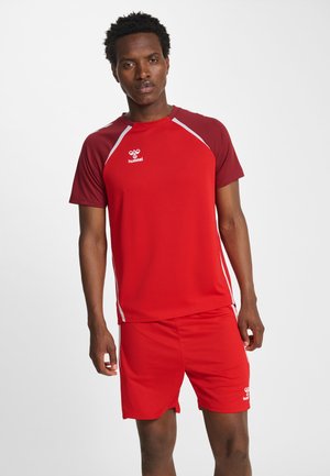 Mann trägt rotes Sportshirt mit kastanienbraunen Ärmeln und passenden roten Shorts, steht vor einfachem weißem Hintergrund.